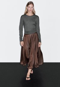 Dlouhá hnědá saténová sukně s plisé, doplněná o přiléhavý šedý top s dlouhým rukávem. Modelka má na sobě černé sandály na rovné podrážce. Jednoduchá estetika, minimální detaily.