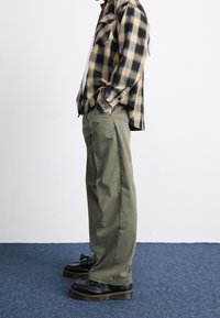 Chemise à carreaux noire et beige avec une coupe décontractée, associée à un pantalon large vert et des chaussures noires à semelles épaisses.