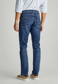 Jeans de mezclilla azul oscuro con un ajuste slim, con dos bolsillos traseros y una etiqueta de cuero en la cinturilla, combinados con zapatos de lona beige.