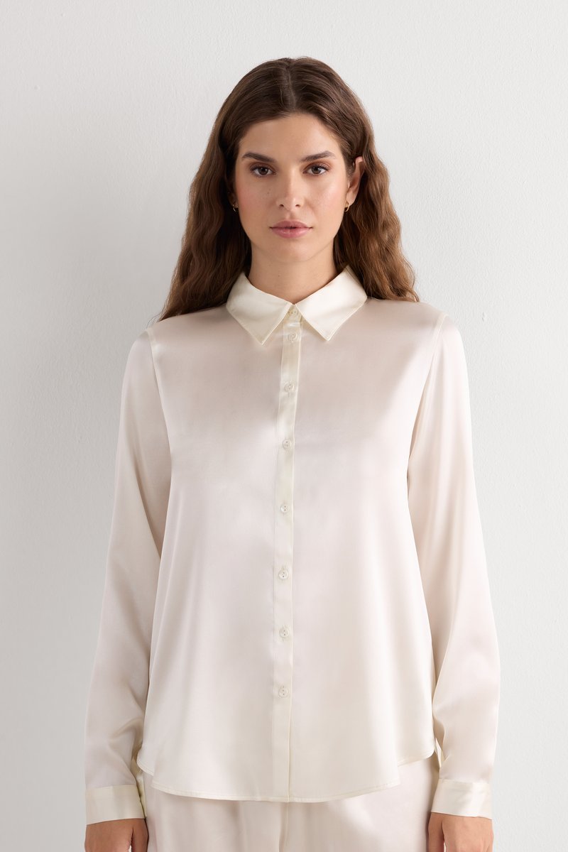 Long-sleeve, light cream blouse made from smooth fabric. Features a classic collar, button front, and a relaxed fit with cuffed sleeves.