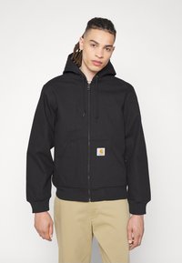 Black zip-up hooded jacket with a front pocket, ribbed cuffs, and hem, featuring a logo patch on the pocket. Smooth fabric texture.