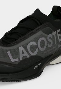 Μαύρο και γκρι αθλητικό παπούτσι Lacoste με μεγάλο λογότυπο στο πλάι, μαύρα κορδόνια και μαλακή σόλα με ετικέτα LSRFOAM.