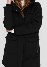 Svart vadderad jacka med blixtlås, två framfickor med klaff och huva. Den har en slät textur med sidofickor med blixtlås.