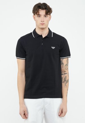 Młody mężczyzna o kręconych brązowych włosach nosi czarną koszulkę polo z białym wykończeniem oraz białe spodnie, na prawej ręce ma widoczne tatuaże.