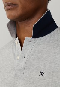 Detail osoby nosiacej svetlosivú polokošeľu s námornícky modrým zdvihnutým golierom a malým logom prekrížených golfových palíc na hrudi.