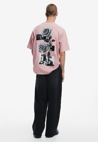 Pink short-sleeve shirt with black graphic prints featuring roses and text on the back. Loose fit with a round neckline and relaxed design.