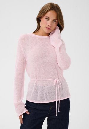 Jeune femme aux cheveux châtain clair portant un pull en maille rose transparent avec un lien à la taille et un jean bleu foncé sur un fond uni.