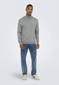 Pulover gri cu guler înalt și mâneci lungi, asortat cu blugi albaștri decolorati și adidași albi, stând pe un fundal simplu.