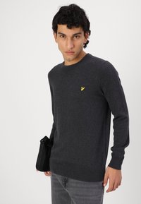Suéter gris oscuro hecho de suave tejido de punto, con cuello redondo y mangas largas. Un pequeño logo amarillo está bordado en el pecho.