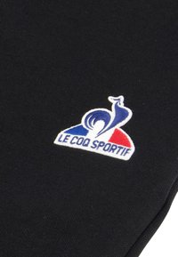 Tissu noir avec un logo brodé d'un coq, coloré en bleu, rouge et blanc, avec le texte "LE COQ SPORTIF" en dessous.