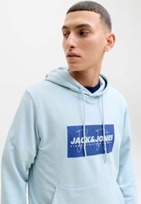 Jeune homme portant un sweat à capuche bleu clair avec le logo "Jack & Jones", regardant sur le côté sur fond blanc uni.