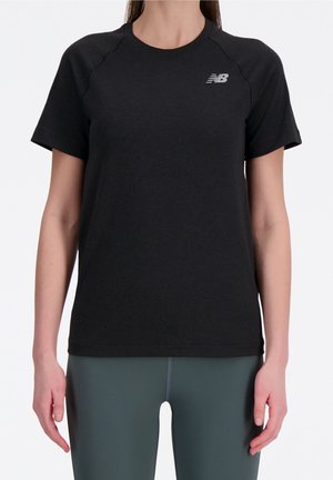 Kvinde iført en simpel sort New Balance t-shirt med korte ærmer og mørkegrønne leggings, stående foran en hvid baggrund.