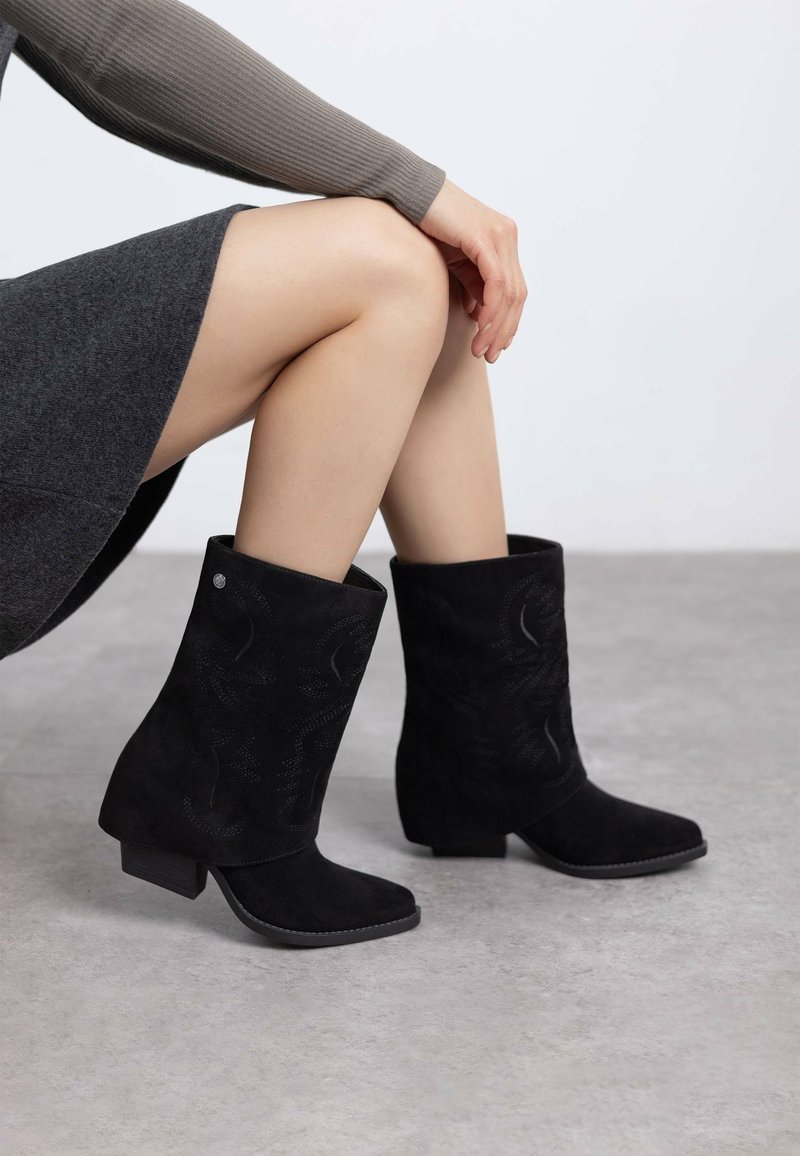 Botas de tobillo de ante negro con tacón cuadrado, que presentan patrones en relieve con remolinos, una puntera redondeada y una textura suave.