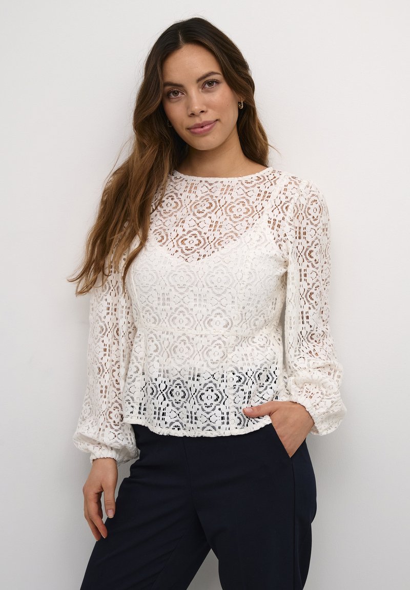 Hvid blondebluse med lange, puffede ærmer og et blomstermønster. Let peplum ved taljen. Sammen med mørke bukser.