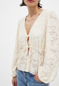 Spitzenbluse in sanftem Elfenbein mit tiefem V-Ausschnitt, Kordelzug in der Taille und puffigen langen Ärmeln. Verfügt über ein florales Muster und eine leichte Textur.