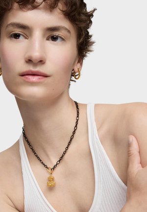 Collana nera a catena con un pendente dorato texturizzato, indossata con una canotta bianca a coste. Dettagli ravvicinati sulla collana e sul décolleté.