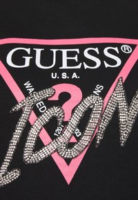 Textil negro con un logo de triángulo rosa que incluye las palabras "GUESS" y "U.S.A." en blanco. El texto "WANTED" y "1203" está adornado con piedras de strás.