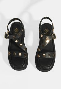 Black leather sandals with a rubber sole, featuring gold-tone studs and crisscross straps. Adjustable buckle closure on the ankle strap.