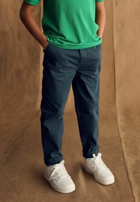 Groene shirt met korte mouwen, donkerblauwe broek en witte sneakers. De broek heeft een rechte snit; de sneakers hebben een geperforeerde structuur en veters.