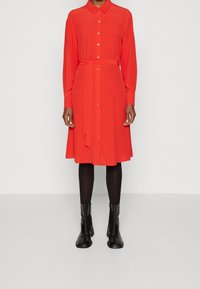 Abito camicia rosso con maniche lunghe, chiusura a bottoni e cintura in vita, realizzato in tessuto liscio. Indossato con calze nere e stivaletti alla caviglia.