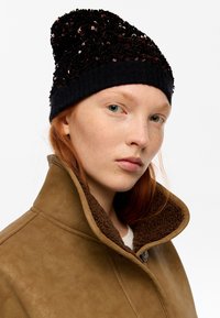 Gorro negro con lentejuelas y borde acanalado, combinado con una chaqueta marrón claro forrada con borrego sintético. Vista en primer plano que enfatiza la textura y los detalles.