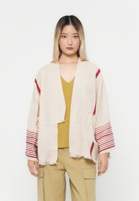 Svijetlosmeđi kimono jakna s crvenim prugama i poderanim rubovima. Nošena preko gornjeg dijela u senfu, u paru s cargo hlačama, pokazujući opušteni kroj.