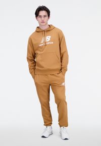 Sudadera marrón con logo blanco, bolsillo delantero y cordón. Pantalones marrones a juego con piernas ajustadas y logo en el muslo. Zapatillas blancas.