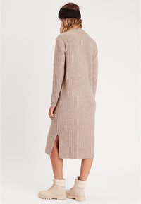 Robe tricotée beige à côtes avec des manches longues, longueur genou et fente latérale. Associée à des bottes beiges à lacets et un bandeau en tissu noir.