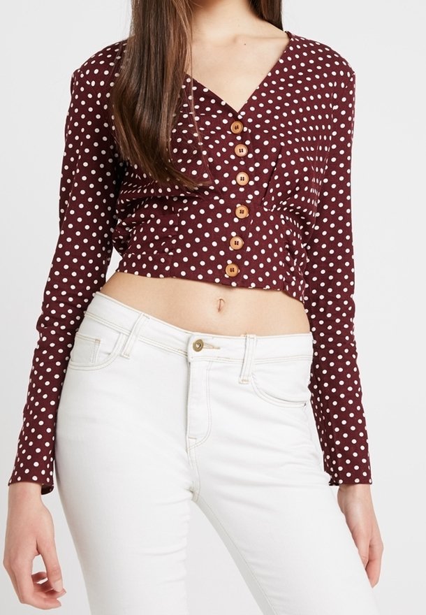 Blouse courte bordeaux à pois blancs, manches longues, col en V et boutons en bois. Associée à un jean blanc taille haute.