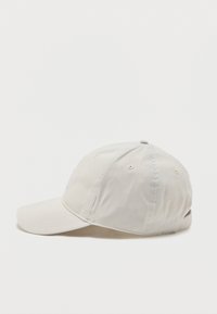 Ανοιχτό μπεζ καπέλο του μπέιζμπολ με κυρτό γείσο, ραμμένα πάνελ και ρυθμιζόμενο λουράκι στο πίσω μέρος, εμφανίζεται από το πλάι σε απλό φόντο.