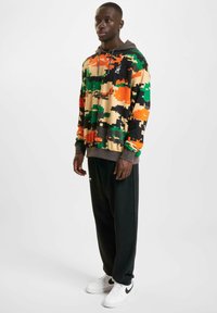 Hooded sweatshirt with pixelated camouflage pattern in orange, green, black, and beige; paired with black trousers and white sneakers.