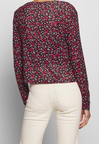 Persona vestida con una blusa negra de manga larga con pequeñas flores rojas y blancas, combinada con pantalones de tiro alto en color blanco roto, vista desde atrás.