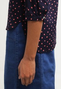 Blouse marine à pois roses, avec des poignets boutonnés, associée à une jupe en denim. Tissu lisse avec une coupe décontractée et une légère ondulation.