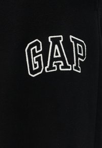 Μαύρο ύφασμα με λευκά περιγράμματα και έντονα κεντημένα γράμματα που σχηματίζουν τη λέξη «GAP» σε καμπυλωτή διάταξη.