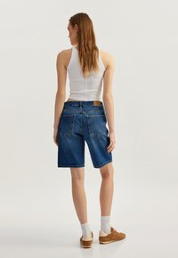 Pantalones cortos de mezclilla azul con cintura alta, cinco bolsillos y un parche de cuero marrón en la parte trasera. Textura suave y corte a la altura de la rodilla.