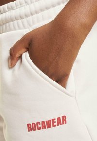White sweatpants with a ribbed waistband and side pocket, featuring a red "ROCAWEAR" logo printed on the thigh area.