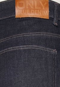 Tmavě modré džínové kalhoty z denimu s koženou etiketou označenou "JEN MODRÝ DENIM", s kontrastním stehováním a dvěma zadními kapsami.