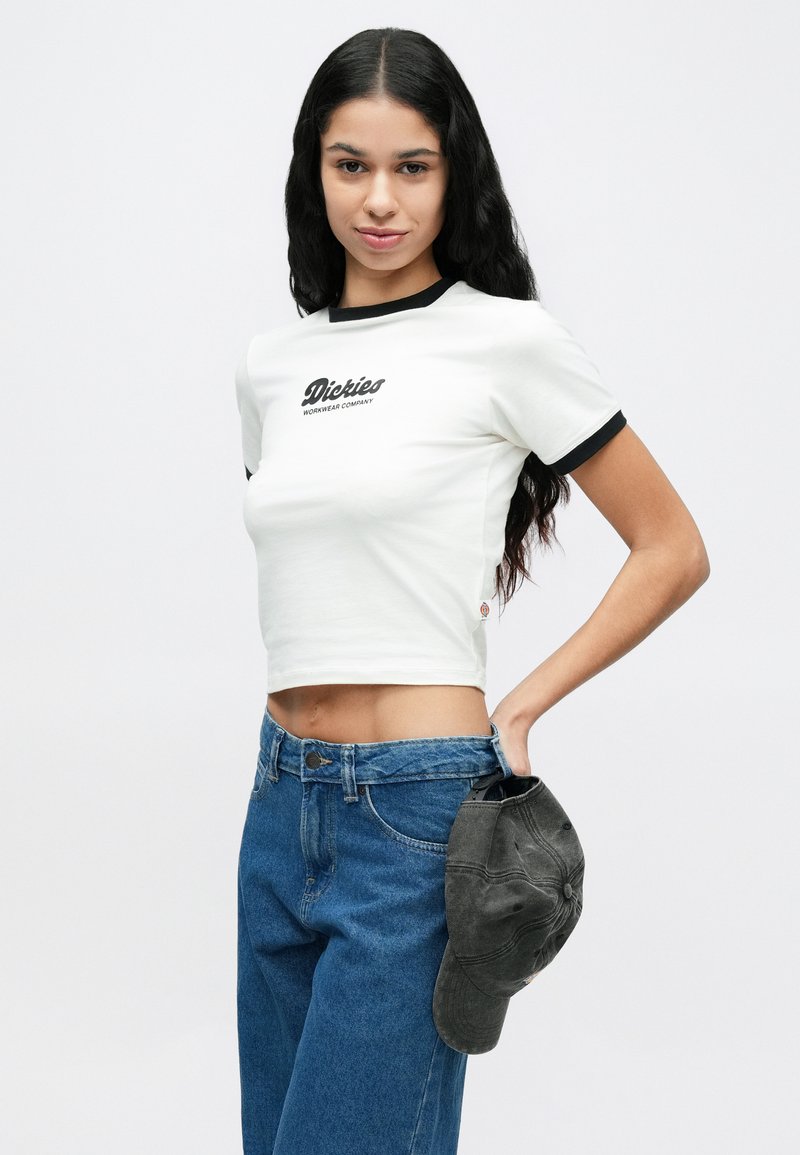 Jeune femme aux longs cheveux bruns portant un t-shirt blanc Dickies court et un jean bleu, tenant une casquette gris foncé à côté d'elle.