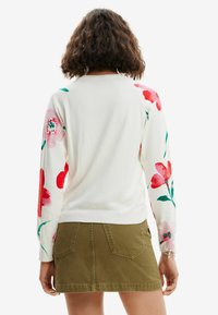 Witte trui met kleurrijke bloemenontwerpen in rood en roze op de mouwen. Gepaard met een groene rok. Zachte brei-textuur, casual pasvorm.