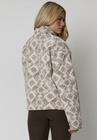 Giacca in fleece beige e marrone con un motivo geometrico, dotata di colletto alto e vestibilità rilassata. Tessuto morbido e testurizzato.