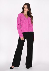 Flauschige rosa Strickjacke mit V-Ausschnitt, Knopfverschluss und verziertem Diamantmuster. Kombiniert mit schwarzen weiten Hosen.