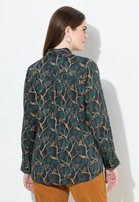 Una donna con lunghi capelli castani indossa una camicia button-up scura con un motivo di uccelli e rami e pantaloni color senape, di fronte a una parete bianca.