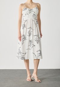 Robe sans manches blanche avec un motif floral noir, buste froncé et ourlet au genou. Assortie à des sandales à brides de couleur claire.