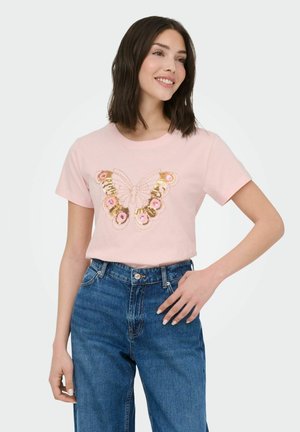 Jeune femme souriante portant un t-shirt rose avec un motif papillon en sequins dorés et roses, assorti à un jean bleu taille haute.