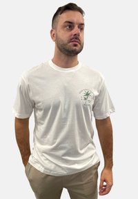 White cotton t-shirt featuring a round neckline, short sleeves, and a green palm tree graphic on the left chest with "One Thousand Palms" text.