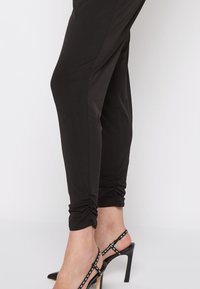 Jambes inférieures portant des leggings noirs froncés et des chaussures à talons hauts noires avec une bride arrière, ornées de coutures blanches, sur un fond uni.
