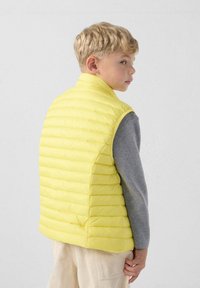 Geel donsloze vest met horizontaal doorgestikt patroon, hoge kraag en een gladde, lichtgewicht textuur, gedragen over een grijze lange mouwen shirt.