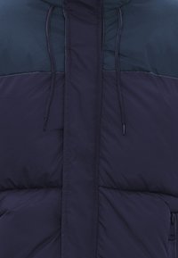 Giacca a vento blu navy con sezione superiore color teal scuro a contrasto, tasche con zip, corde di regolazione nel cappuccio e texture trapuntata.