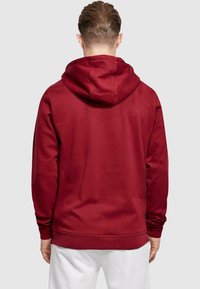 Röd sweatshirt med huva, med en mjuk yta och ribbade ärmslut. Bakifrån syns inga synliga logotyper eller mönster.