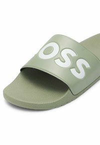 Sandale slide verte à bout ouvert avec semelle texturée et large bride portant de grandes lettres blanches "O", "S", "S".